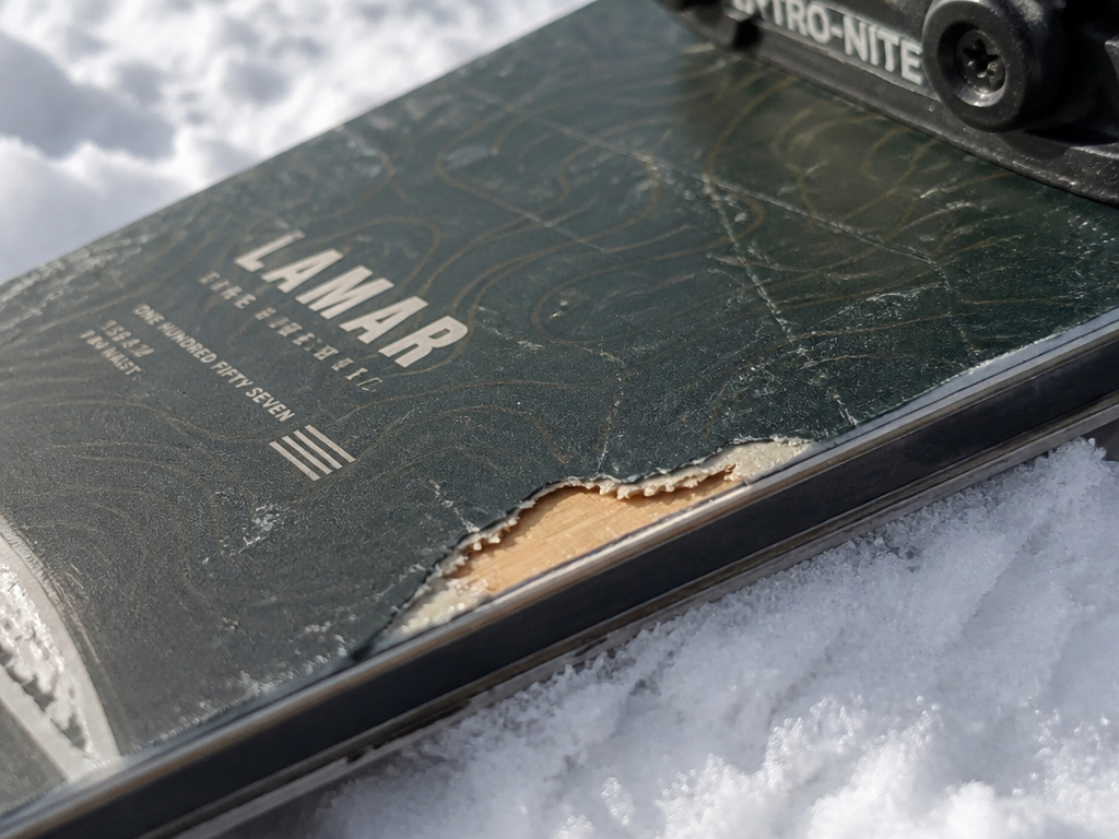Close-up of chipped topsheet damage on a used Lamar snowboard.